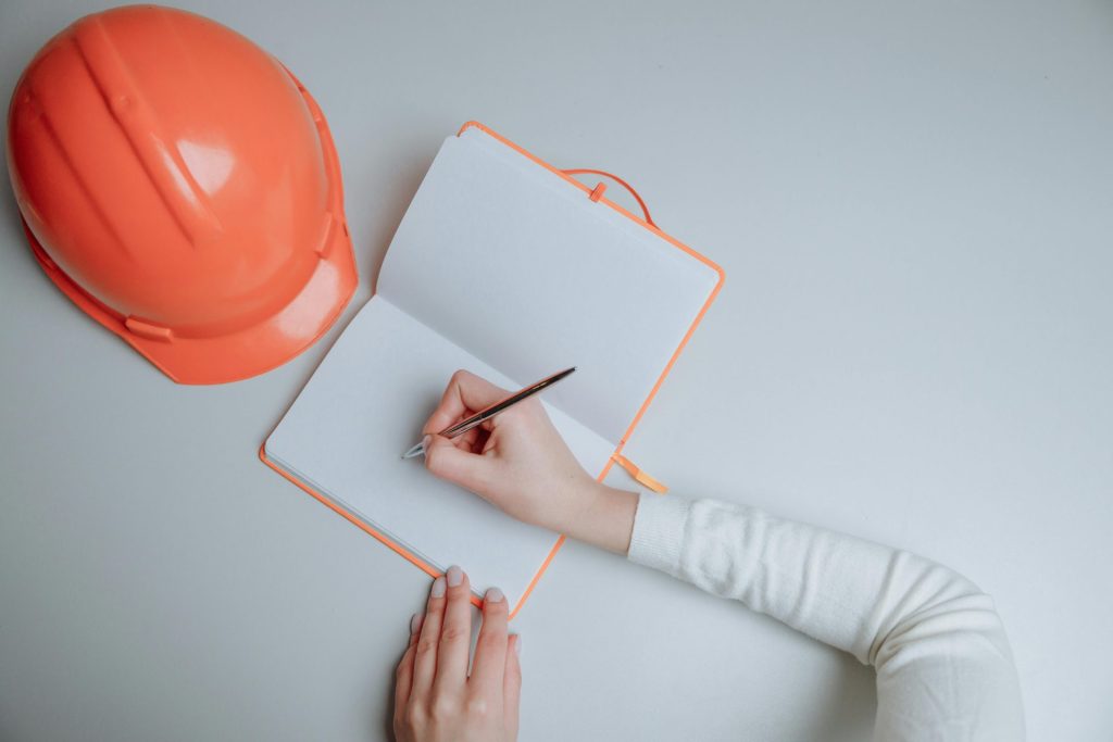 Draufsicht auf eine Person, die neben einem orangefarbenen Schutzhelm vor weißem Hintergrund in ein Notizbuch schreibt.